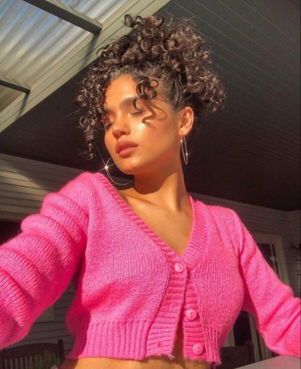 A woman with naturally curly hair pulled up in a high curly ponytail, wearing a vibrant hot pink cropped cardigan and large hoop earrings, soaking in the golden hour light.