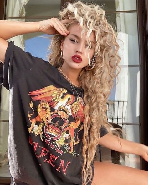 Woman with voluminous, tight blonde curls wearing a Queen band t-shirt, posing with a hand on her forehead