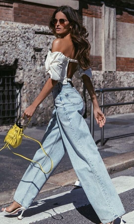 A woman walks confidently in light-wash baggy jeans paired with an off-the-shoulder white crop top, white heels, and a small yellow handbag.