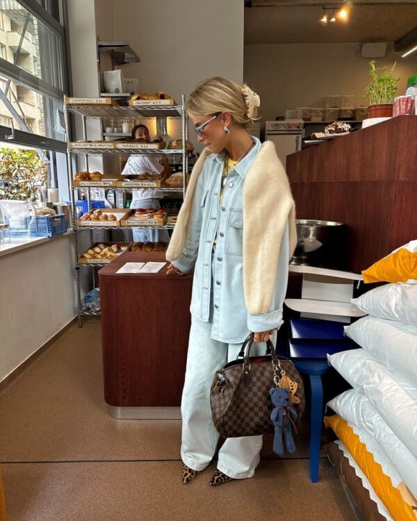 A woman wearing a light-wash denim set with a cream sweater draped over her shoulders, holding a designer handbag.