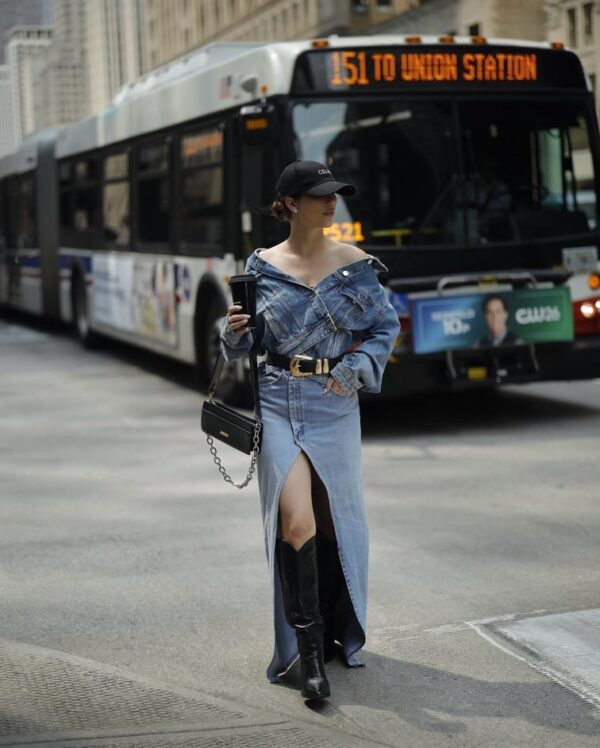 A woman is wearing an off-shoulder denim shirt with a side cut-out, black boots, and a baseball hat.