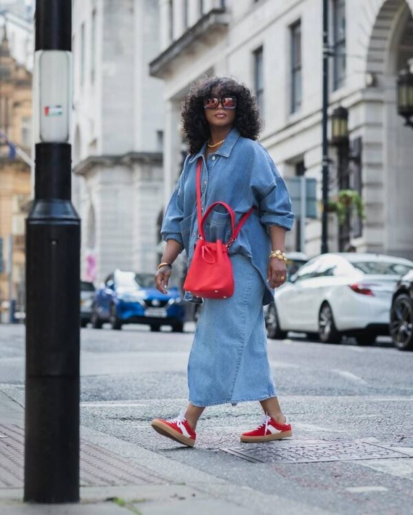 Woman in a denim maxi skirt, oversized shirt, red handbag, and matching red sneakers.