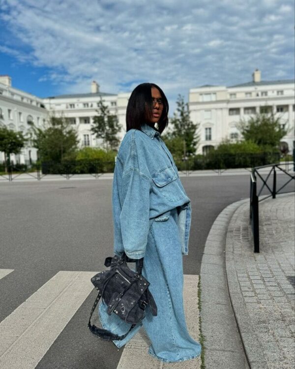 Woman in oversized denim layers, holding a rugged black leather bag.