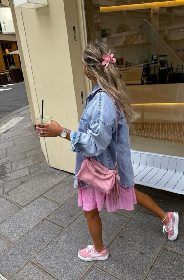 Woman wearing a pink dress, oversized denim jacket, pink sneakers, and a pink flower hair clip while holding an iced coffee