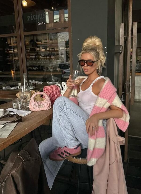 Woman wearing light wash jeans, a white tank top, a pink and white striped sweater, and pink sneakers, sitting outside a café with a glass of wine