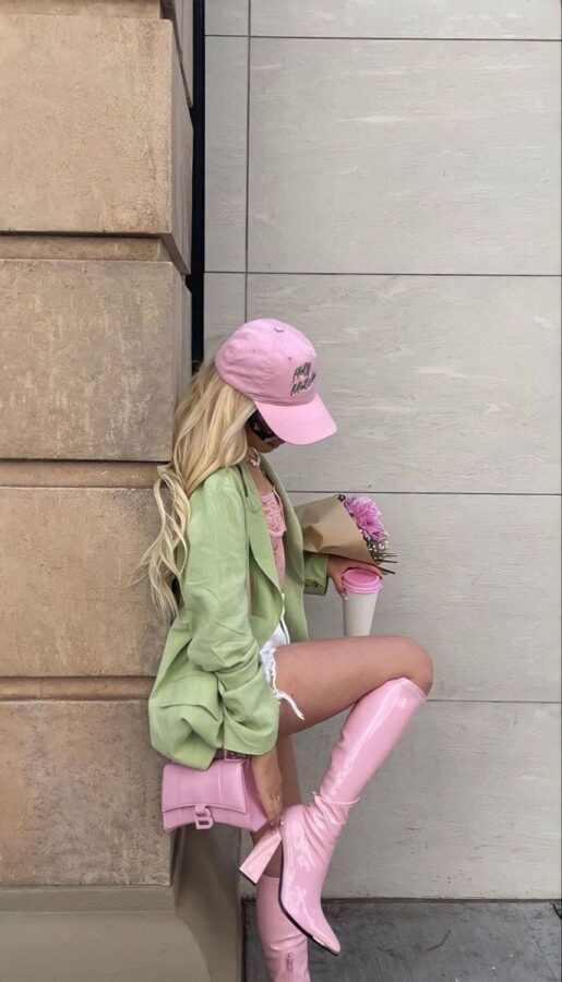 Stylish woman in pastel green blazer, pink knee-high boots, pink bag, and pink cap holding flowers and coffee