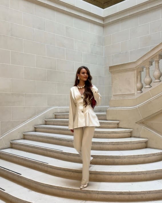 A woman wearing an ivory satin blazer and matching trousers while descending elegant stairs.