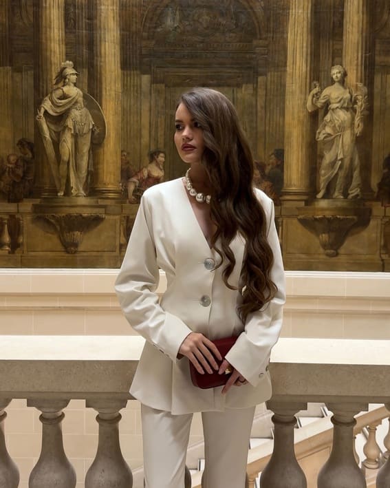 A woman in an ivory suit with a large pearl necklace is in front of a classical backdrop.