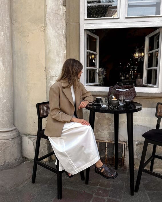 A woman in a tan blazer and white midi dress is sitting at a café table.