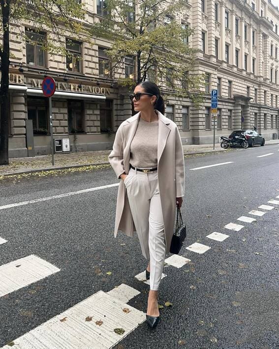 A woman in a beige overcoat and white trousers walks on a city street.