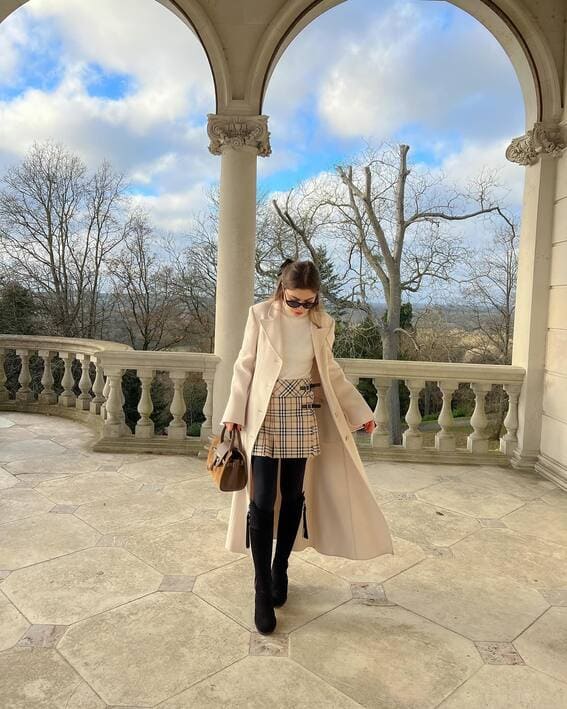 A woman in a cream coat, plaid skirt, and high boots stands under an archway.