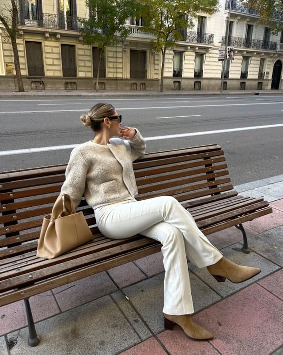 A woman in a cream sweater, white pants, and tan boots sits on a bench.