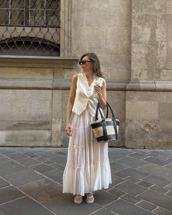 Woman in a long white dress and vest with a straw bag.