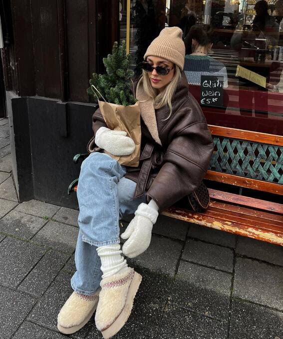 A woman in a layering winter outfit wears a warm coat, blue jeans, fuzzy Ugg slippers, and a beanie.