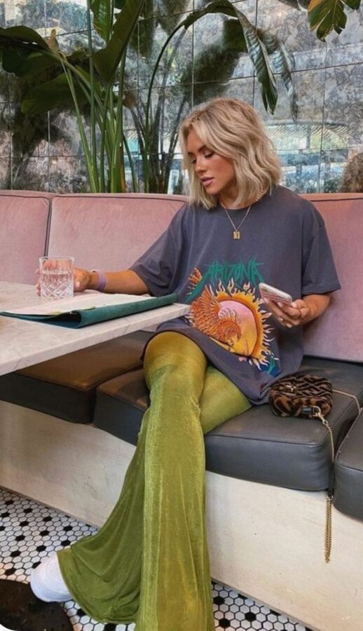 Woman wearing a graphic oversized tee, green velvet flared pants, and white sneakers, sitting at a café with a drink and menu