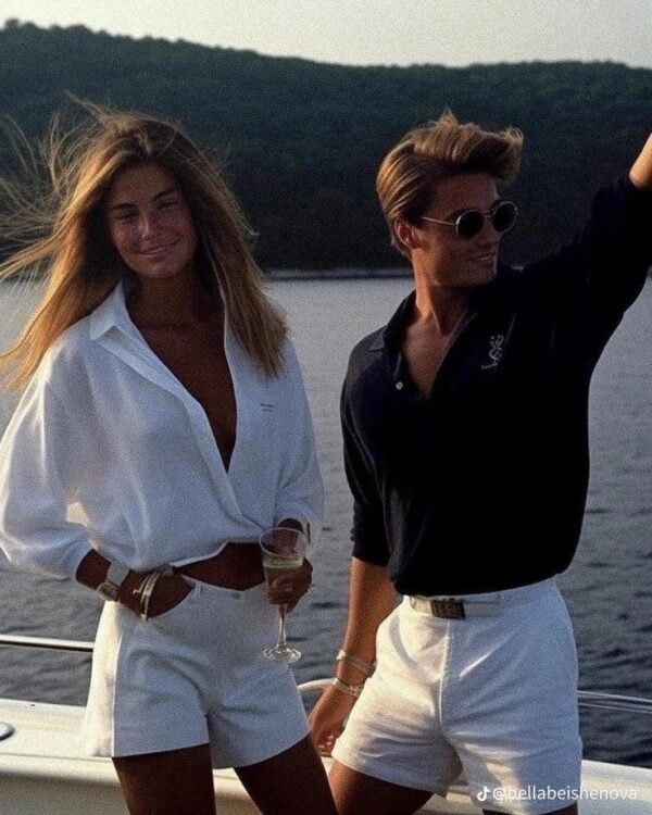 A woman in white high-waisted shorts and an open white button-up shirt standing next to a man in a navy polo and white shorts, both on a boat at sunset.