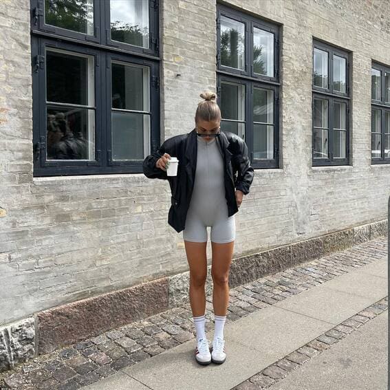 This relaxed and sporty look features an oversized grey romper, a black leather jacket, white sneakers, and socks.