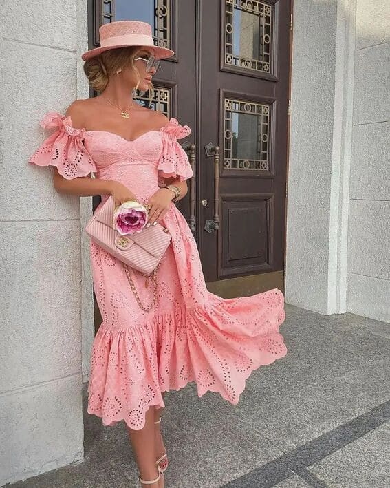 A woman in a pink eyelet dress with a matching hat, carrying a Chanel bag and standing against an ornate doorway.