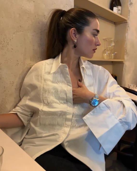 A woman in an oversized white shirt with statement sleeves, accessorized with a silver watch and drop earrings, sits at a restaurant.