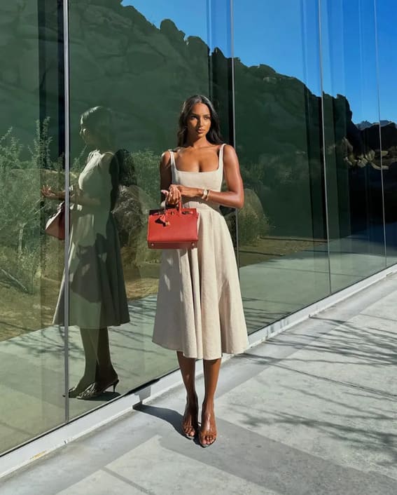 A woman in a beige midi dress holding a red handbag stands in front of a modern glass building.