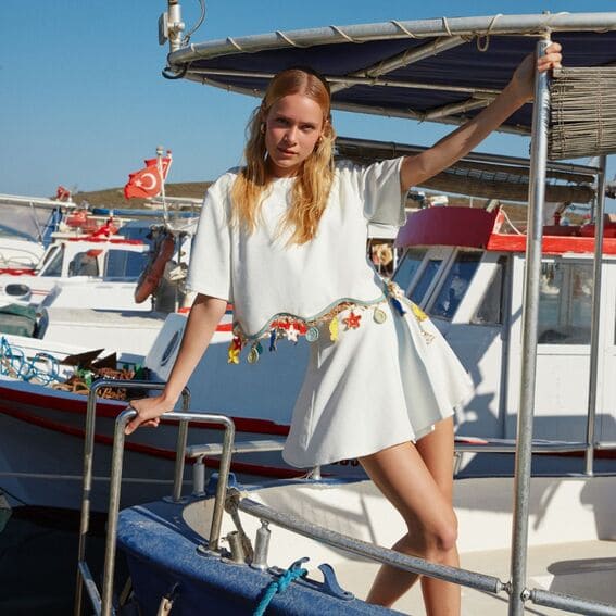 A woman in a white, cropped outfit with colorful embroidered details stands on a boat docked by the marina.