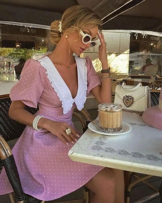 A woman in a pink polka dot dress with a white collar, accessorized with white sunglasses, pearl jewelry, and a structured white handbag, enjoying coffee at a café.