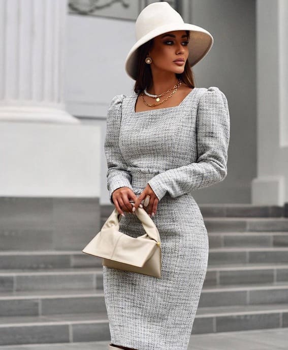 A woman in a tailored grey tweed dress, carrying a structured cream handbag and a wide-brimmed hat, stands in front of elegant stone steps.