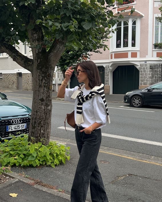 A woman in a white t-shirt, black jeans, and sunglasses, with a striped sweater draped over her shoulders, walks down a quiet street.