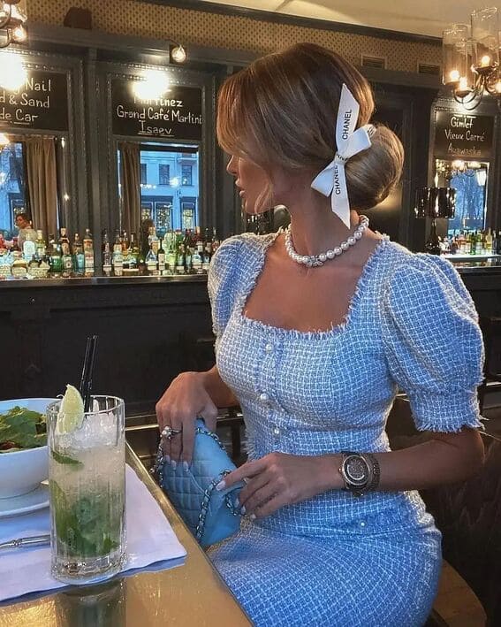 A woman in a light blue tweed dress with pearl detailing, wearing a Chanel ribbon in her updo, is seated at a bar.