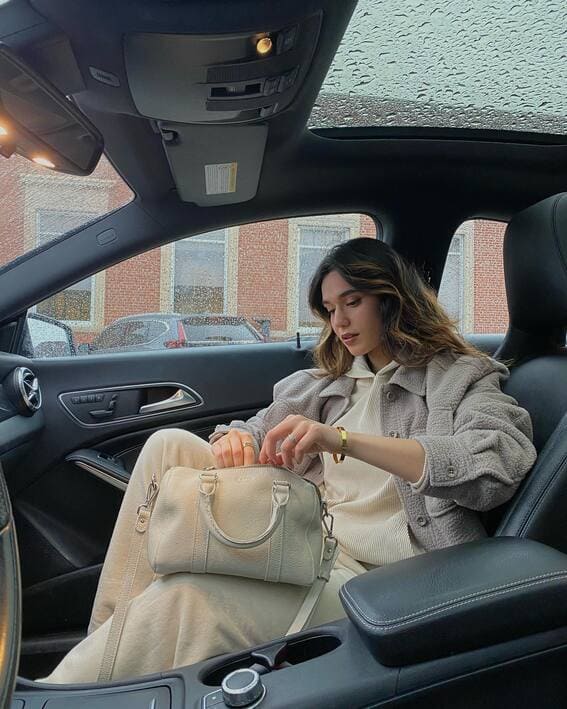 A cozy monochrome beige outfit, including a wool jacket and a cream bag, is worn inside a car on an overcast day. 