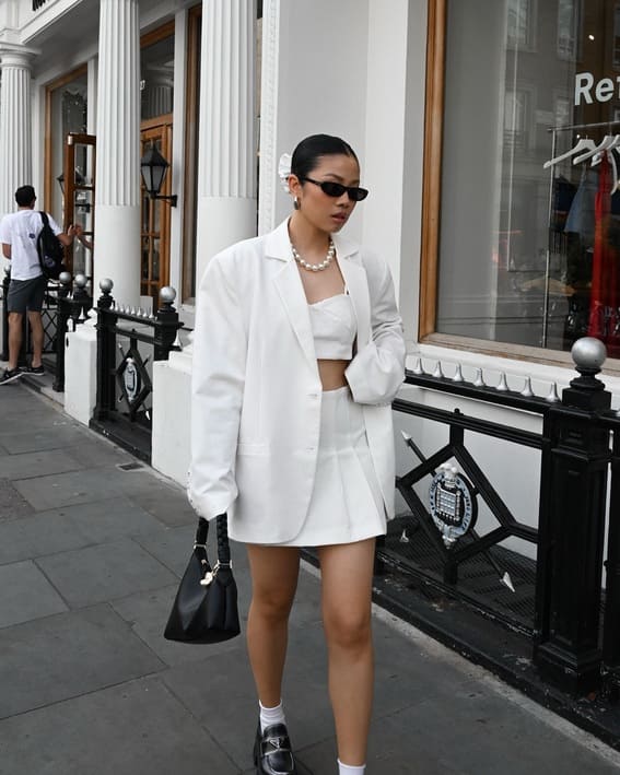 Stylish all-white monochrome outfit with an oversized blazer, mini skirt, and loafers on a city sidewalk.