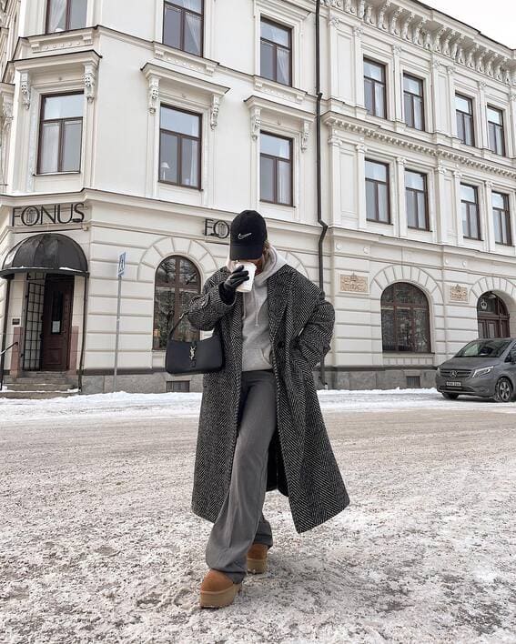 Monochrome winter outfit with a herringbone coat, gray trousers, and platform boots in a snowy cityscape. 