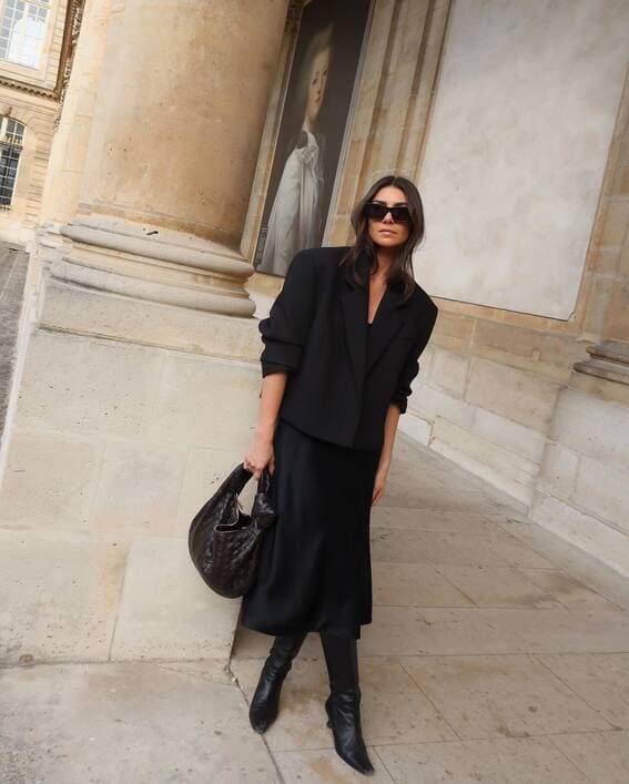 Elegant monochrome black outfit with a structured blazer, satin skirt, and knee-high boots. 