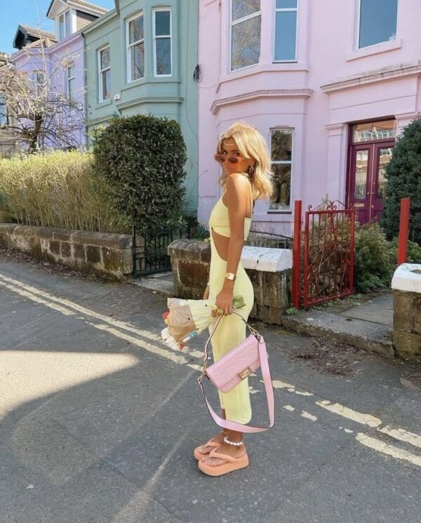 Woman wearing a pastel yellow matching set with a pink bag and peach sandals, holding flowers on a colorful street