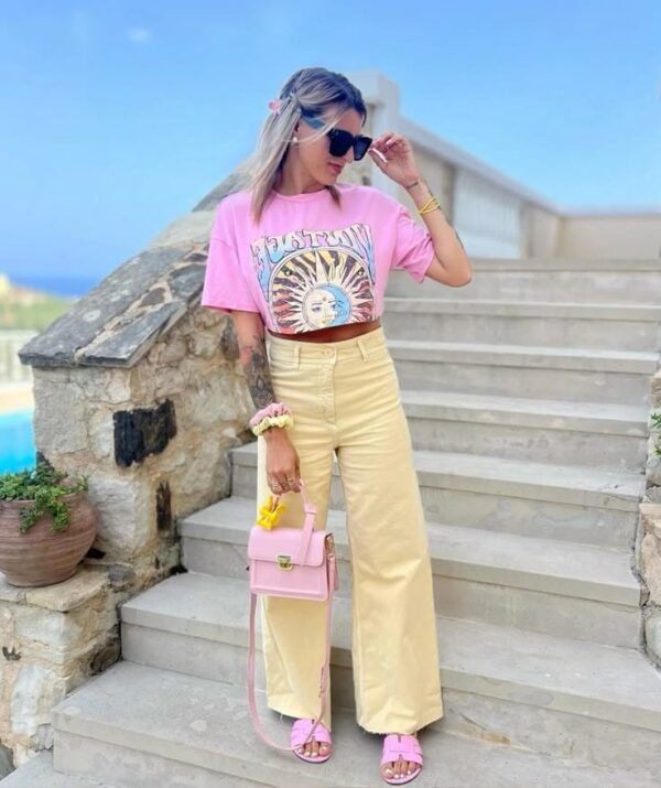 Woman wearing a pink graphic crop top, butter yellow wide-leg pants, pink sandals, and a pastel pink handbag with sunny accessories.