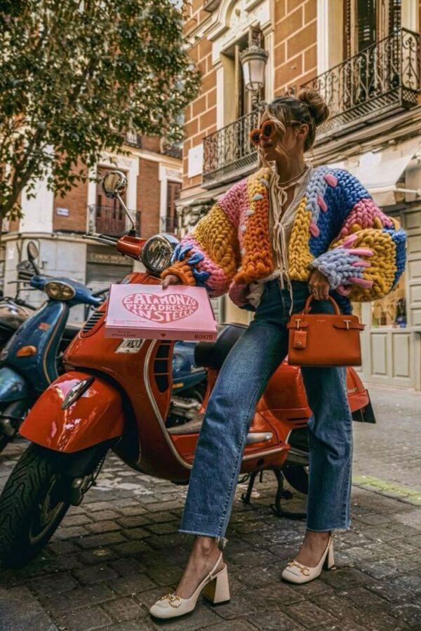 Woman wearing a chunky colorful knit sweater, straight-leg jeans, white heels, and carrying an orange handbag