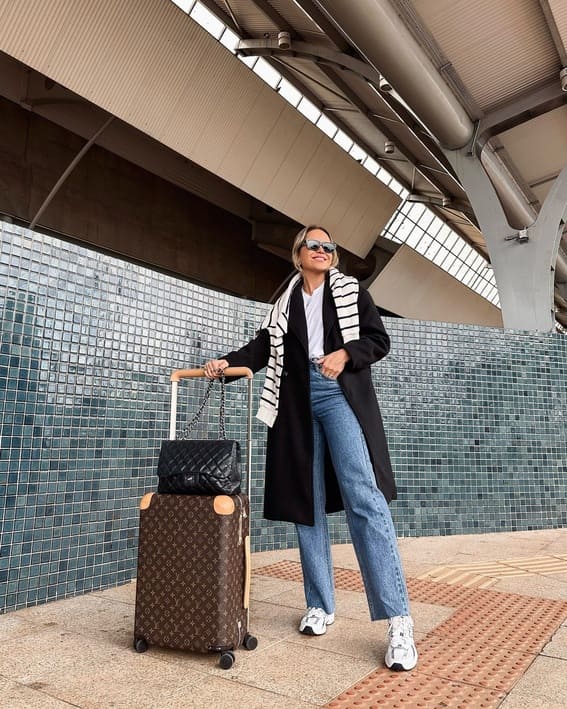 A woman in a long black coat, white sneakers, and a striped scarf pulled a Louis Vuitton suitcase at the airport.