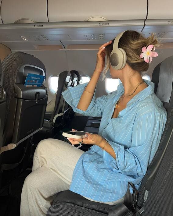 A woman in a blue-striped shirt and beige linen pants sits on a plane with headphones and a plumeria hair clip.