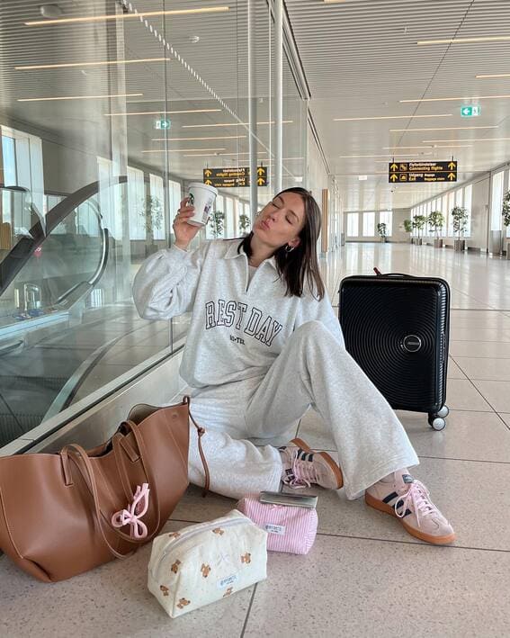 A woman in a grey tracksuit, pink Adidas sneakers, and a brown leather tote is sitting at the airport.