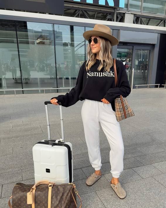 A woman in a black sweatshirt, white joggers, and a beige wide-brimmed hat stands at the airport.