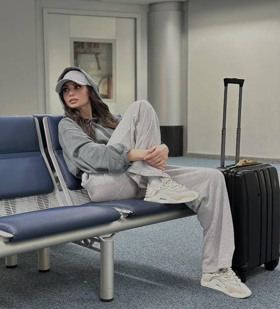 A woman in grey sweatpants and a sweatshirt sits at an airport terminal with a black suitcase.