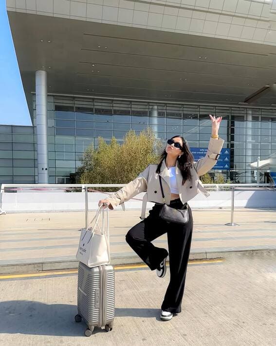 A woman in a cropped beige jacket, black wide-leg trousers, and sneakers stands by a suitcase.