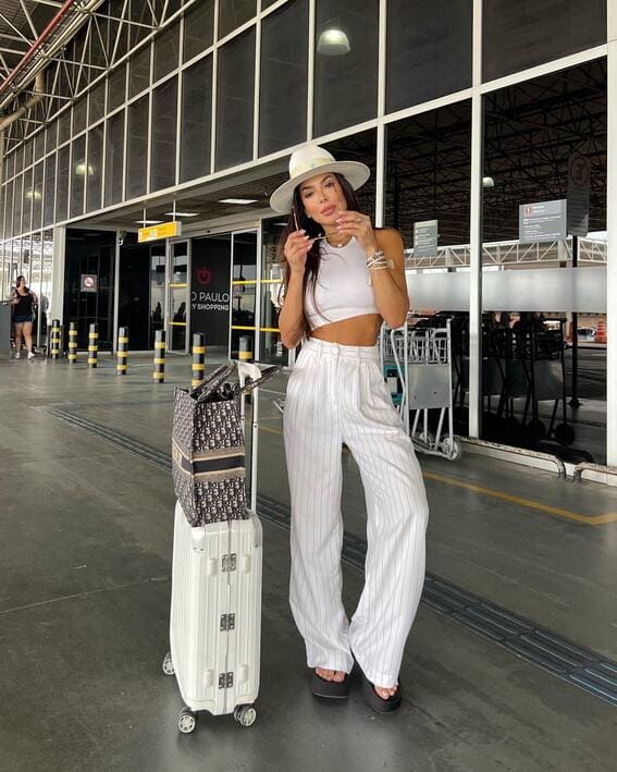 A woman in white pinstriped trousers, a cropped tank, and a wide-brimmed hat standing outside the airport with a suitcase.