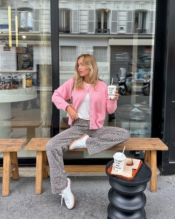 A woman wearing a pink cardigan and leopard-print pants is seated in a cafe and enjoying coffee.