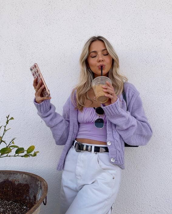 A female is wearing a lilac crop top, matching cardigan, white high-waisted jeans, and an elastic belt. She is sipping an iced cup of coffee against a white backdrop.