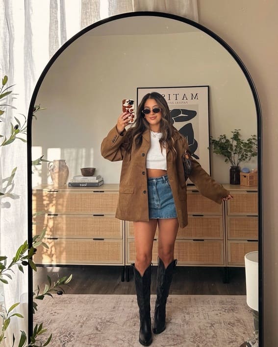 A woman dressed in a cropped white shirt with a mini skirt of denim, an oversized tan jacket, and black boots. An image of herself in a mirror.