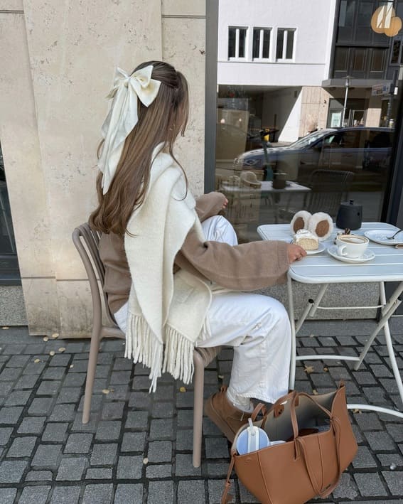 A woman sporting a cream scarf, white pants, a beige coat, and a massive cream bow tied in her hair. She is seated at a cafe table outside.