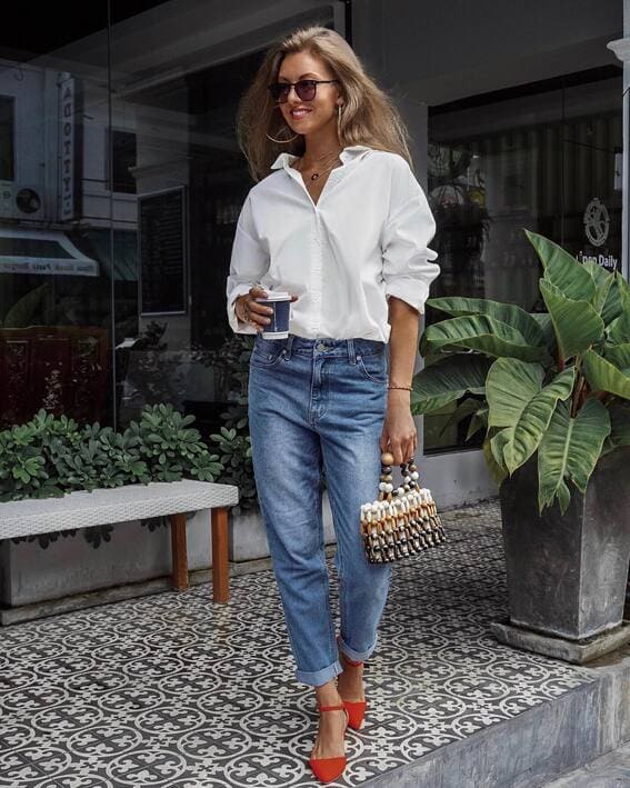 A female dressed in a crisp white button-down t-shirt, light-wash jeans, pointed red heels, and a handbag with beads and a cup of coffee in front of a cafe.