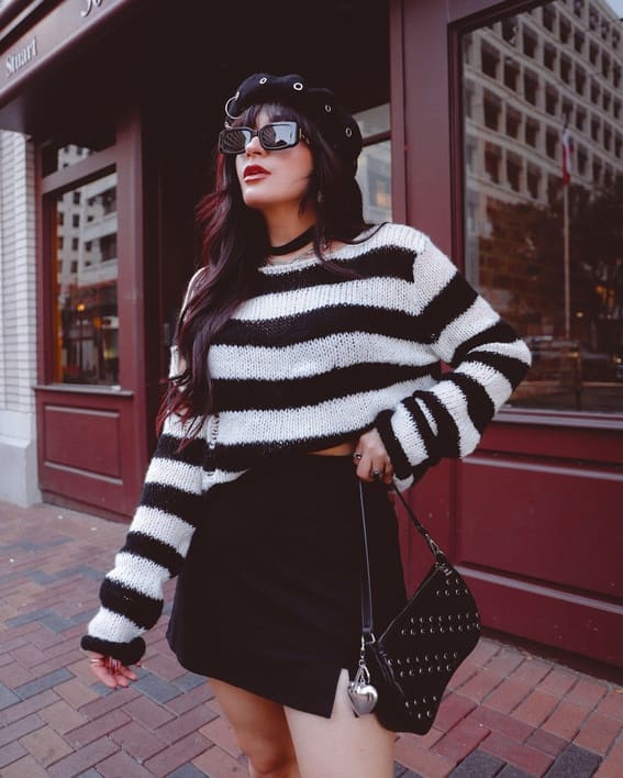 A woman wearing a white and black uni-colored shirt, black mini skirt beret, and dark sunglasses while carrying a studded bag outside a café.