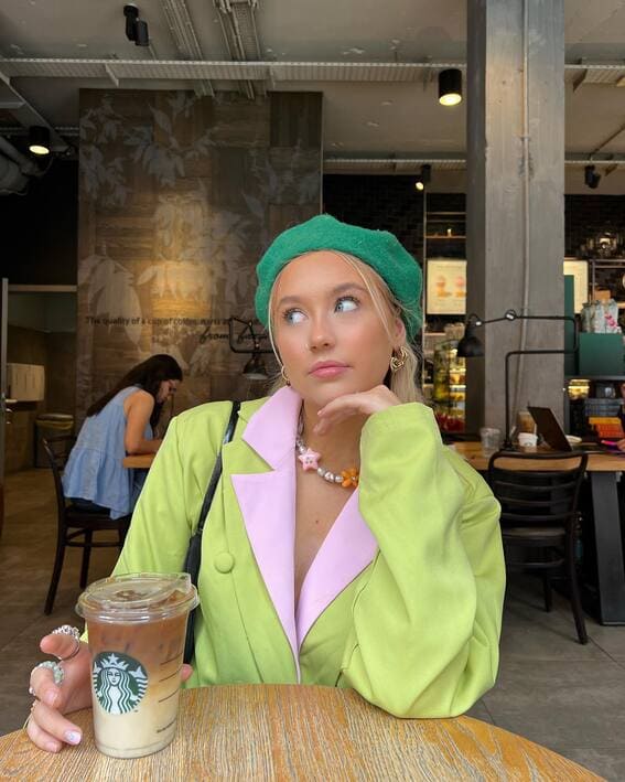 A female wearing a lime green blazer with rose-colored lapels and a green beret sits in a café with a cup of iced coffee.